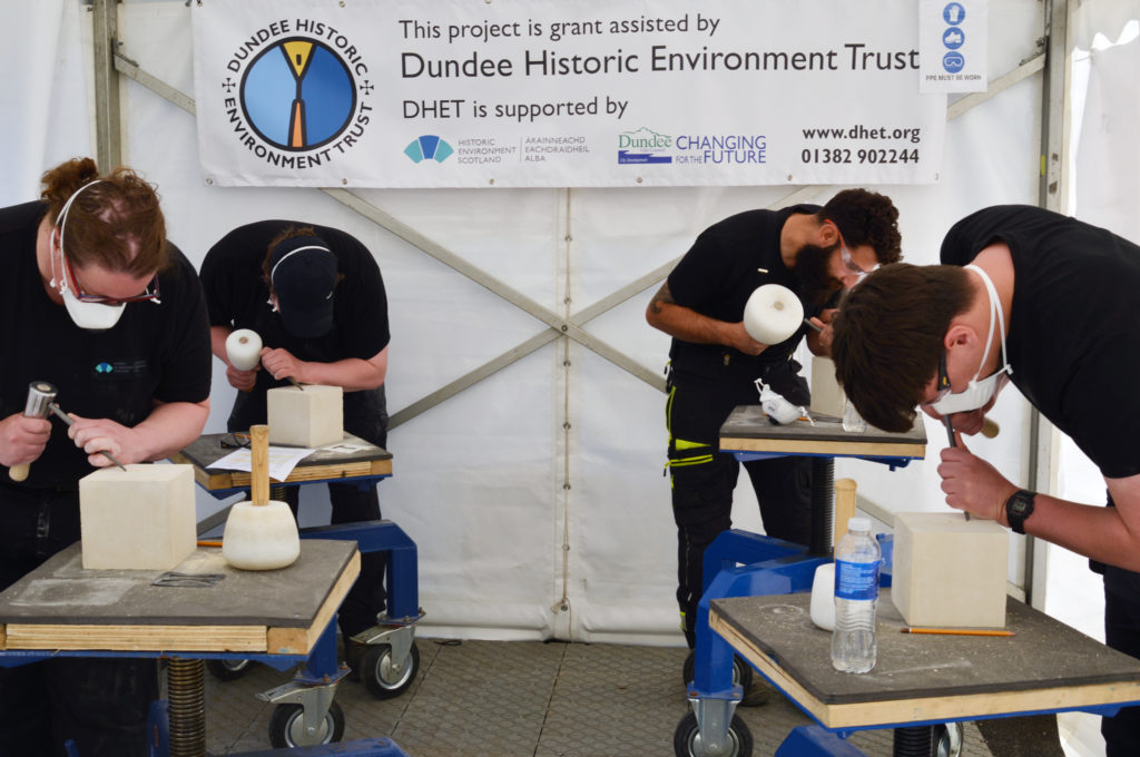 Four people work on stone carvings inside a marquee. They wear goggles and masks.