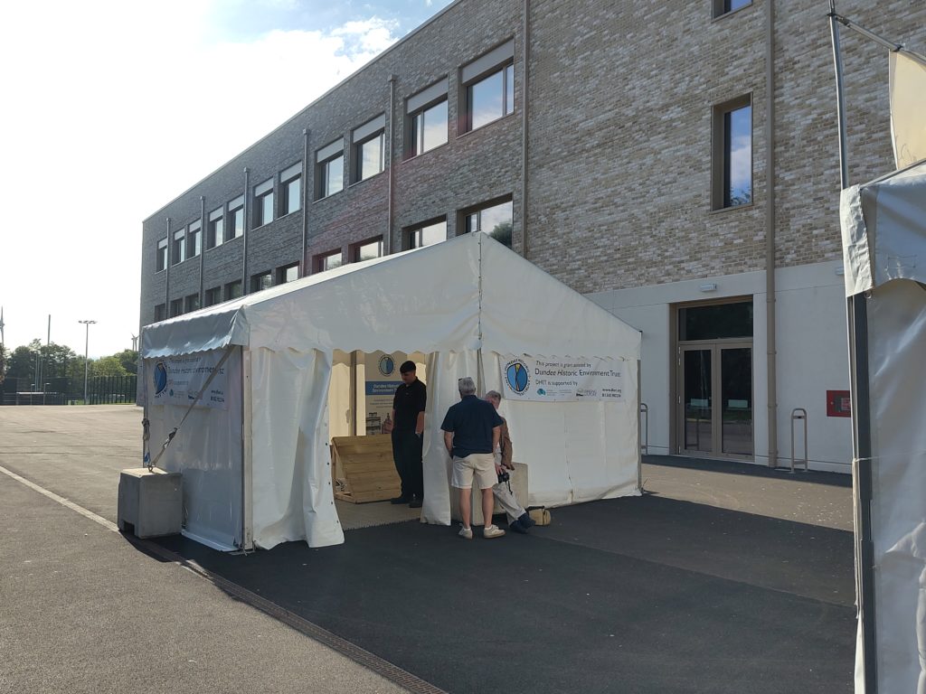 A white marquee outside a brick building on a sunny morning. Two people are outside the marquee and one can be seen through the doorway